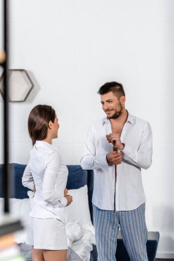 cheerful handsome boyfriend buttoning cuff and looking at girlfriend buttoning her shirt in weekday morning in bedroom, gender equality concept clipart