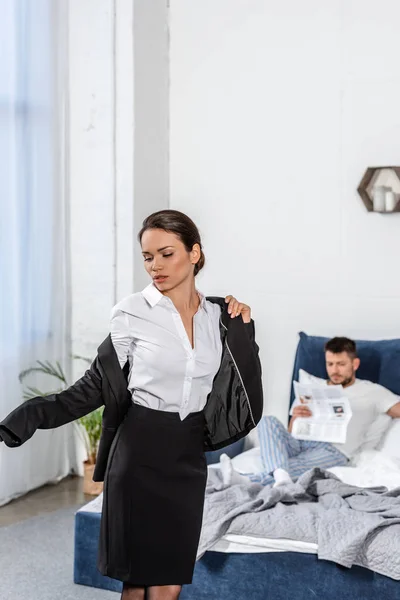 girlfriend wearing jacket and boyfriend in pajamas reading newspaper in bedroom in morning at weekday, gender equality concept