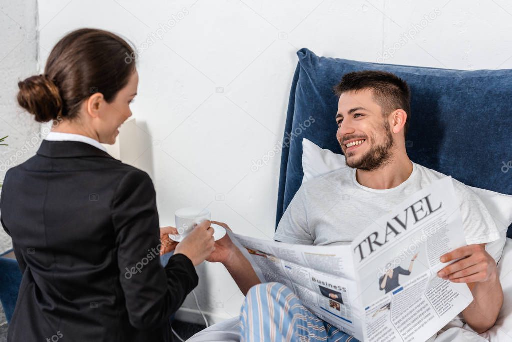 girlfriend in suit giving cup of coffee to smiling boyfriend in pajamas in bedroom in morning, social role concept