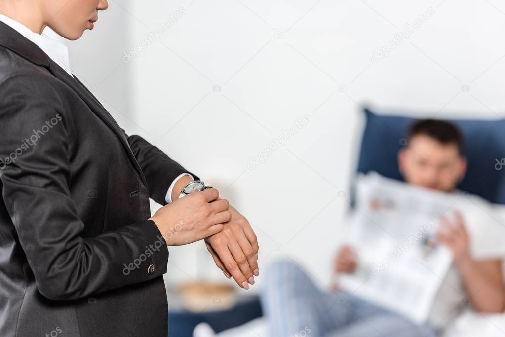 Cropped image of girlfriend in suit checking time at wristwatch and boyfriend in pajamas reading newspaper in bedroom in morning, gender equality concept