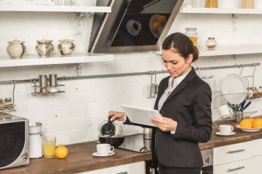 attractive woman pouring coffee into cup and looking at tablet in morning at kitchen clipart