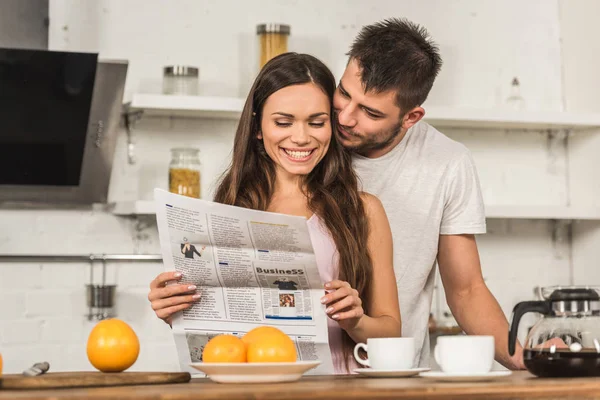 Smiling Girlfriend Reading Newspaper Boyfriend Hugging Her Back Morning Kitchen — Stock Photo, Image