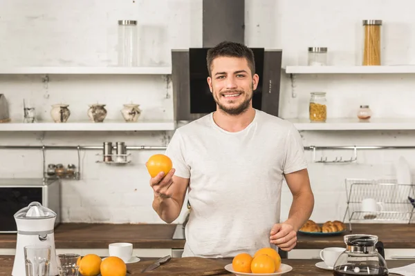 ハンサムな笑みを浮かべて男オレンジを押しながらキッチンで朝のカメラを見て  — 無料ストックフォト
