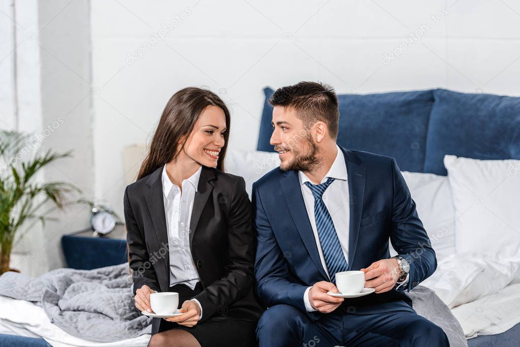 smiling girlfriend and boyfriend in suits sitting on bed and holding cups of coffee in morning at home, gender equality concept