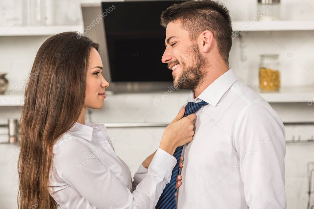 portrait of girlfriend tying smiling boyfriend tie in morning at kitchen, sexism concept