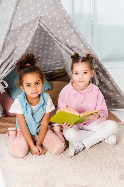 kitap ve kameraya gülümseyen halı üzerinde oturan sevimli çok ırklı çocuklar yüksek açılı görünüş