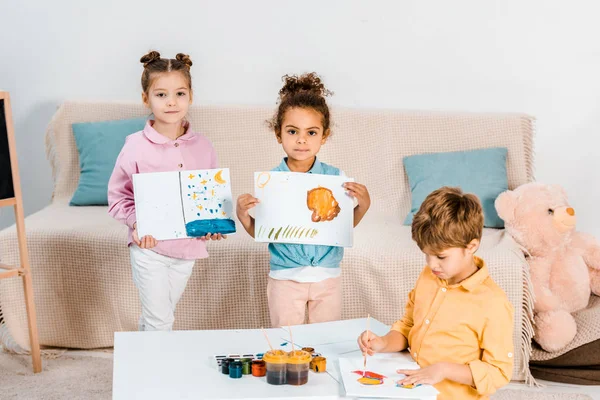 Lindos Niños Multiétnicos Sosteniendo Imágenes Mirando Cámara Mientras Pintan Juntos —  Fotos de Stock