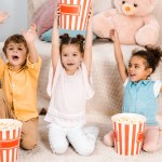 Roztomilý šťastné děti sedí na koberci a drží boxy s popcorn