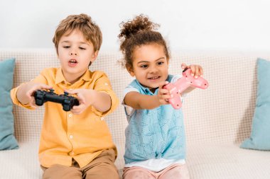 Kanepede oturan ve joystick ile bilgisayar oyunu oynamak sevimli duygusal çok ırklı çocuklar