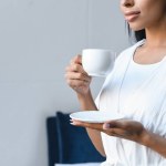 Обрізане зображення змішаної гонки дівчини в білому халаті, що тримає чашку кави вранці в спальні