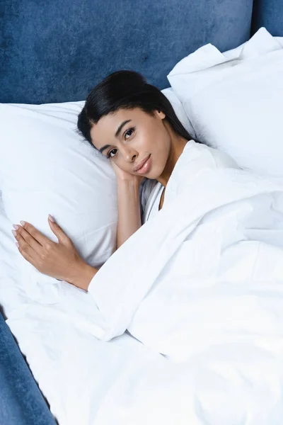 美丽的混血女孩在白色长袍的高角度的看法躺在床上 在早晨看着相机 — 免费的图库照片