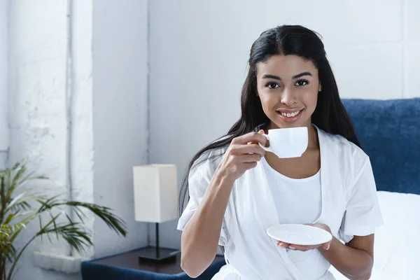 微笑美丽的混血女孩在白色长袍喝咖啡 看着相机在早上的卧室 — 免费的图库照片