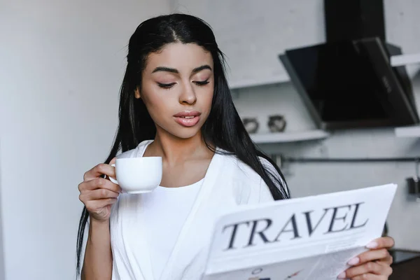 有吸引力的混血女孩在白色长袍举行一杯咖啡和阅读旅游报纸在早上在厨房 — 免费的图库照片