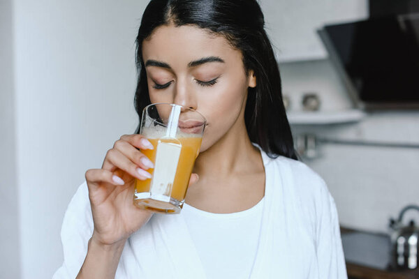 beautiful mixed race girl in white robe drinking fresh orange juice in morning in kitchen with closed eyes