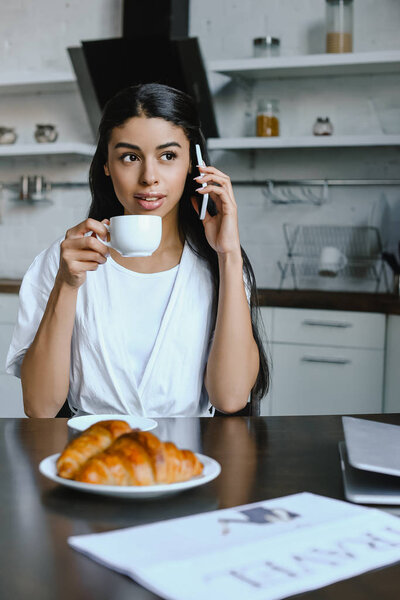 attractive mixed race girl in white robe talking by smartphone in morning in kitchen and drinking coffee