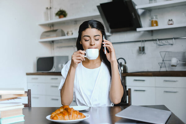 beautiful mixed race girl in white robe talking by smartphone in morning in kitchen and drinking coffee with closed eyes