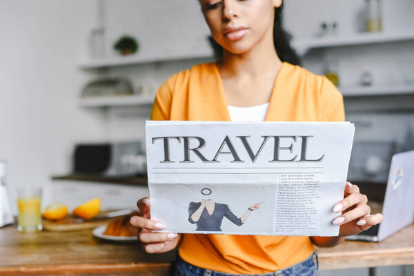 selective focus of beautiful mixed race girl in orange shirt holding travel newspaper in kitchen