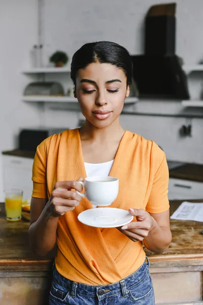 有吸引力的混血女孩在橙色衬衫举行一杯咖啡和盘子在厨房里 — 图库照片