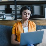 Красивая девушка смешанной расы в оранжевой рубашке слушает музыку и использует ноутбук в гостиной Красивая девушка смешанной расы в оранжевой рубашке слушает музыку и использует ноутбук в гостиной