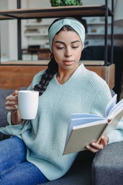 attractive mixed race girl in turquoise sweater and headband sitting on sofa and holding cup of tea and reading book in living room clipart