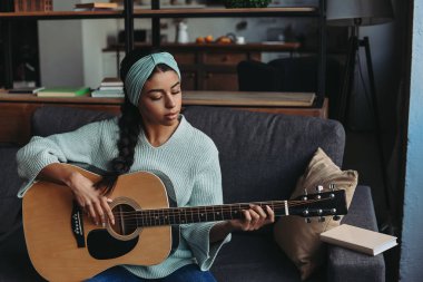 güzel karışık yarış kız turkuaz kazak ve evde kanepede akustik gitar çalmak kafa bandı