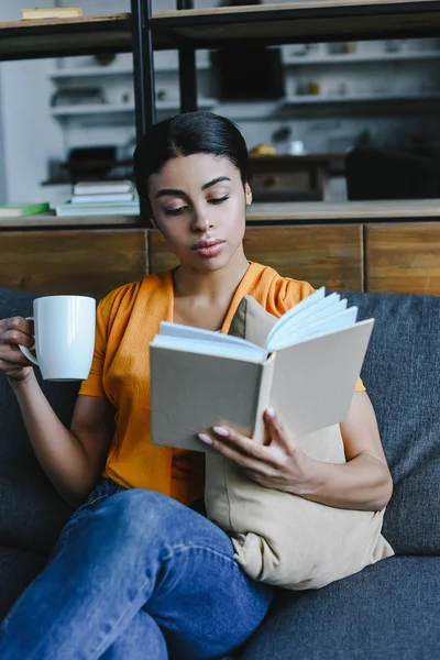 美丽的混血女孩在橙色衬衫拿着一杯茶和阅读书籍在家里的沙发上 — 图库照片