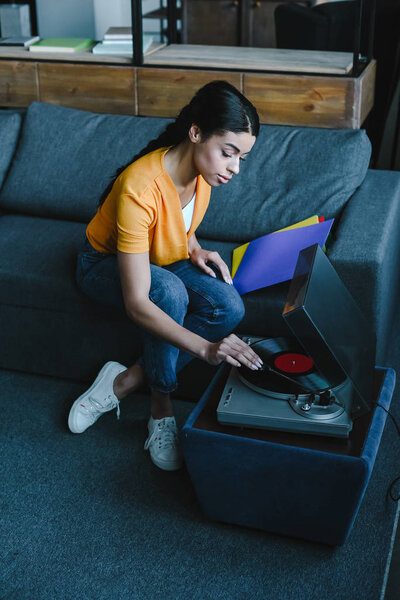 high angle view of beautiful mixed race girl in orange shirt turning on gramophone with retro vinyl at home