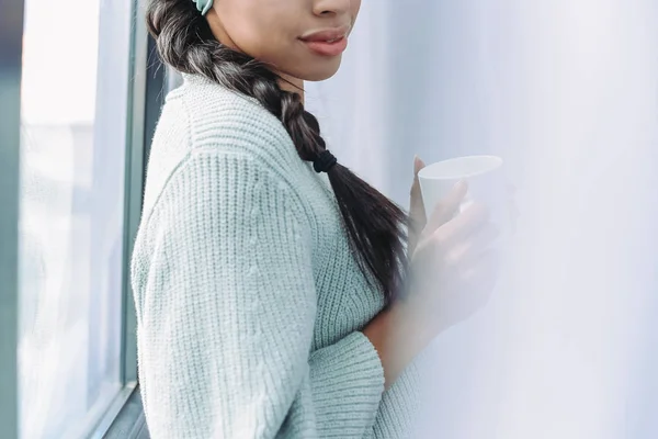 家にお茶のカップを保持している青緑色のセーターで混血の少女のトリミングされた画像 — ストック写真