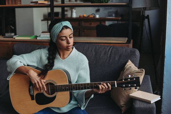 beautiful mixed race girl in turquoise sweater and headband playing acoustic guitar on sofa at home