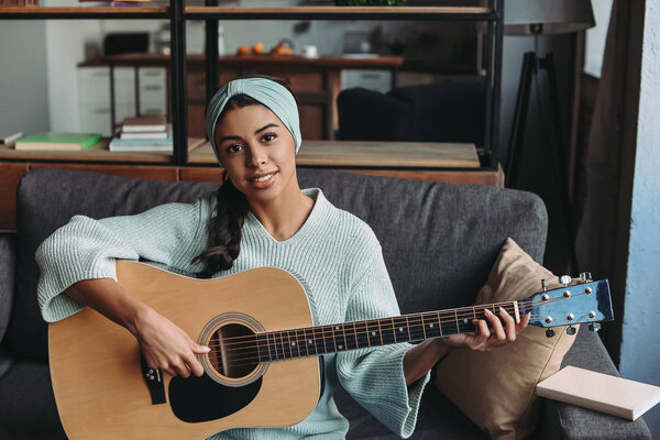 attractive mixed race girl in turquoise sweater and headband playing acoustic guitar on sofa at home and looking at camera