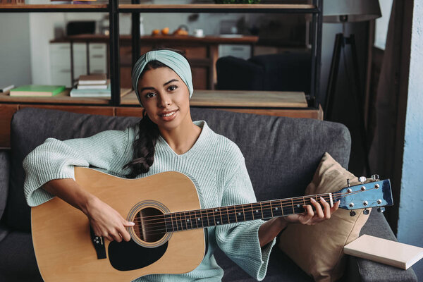 привлекательная девушка смешанной расы в бирюзовом свитере и ободок, играющая на акустической гитаре на диване дома и смотрящая в камеру
