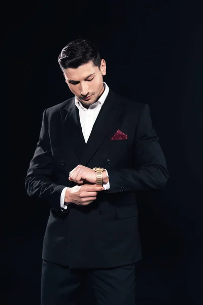 focused man in suit putting on watch isolated on black