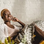 Cheerful african american woman lying on yellow sofa under blanket