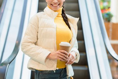 cropped view of smiling girl going down on escalator and holding paper cup clipart