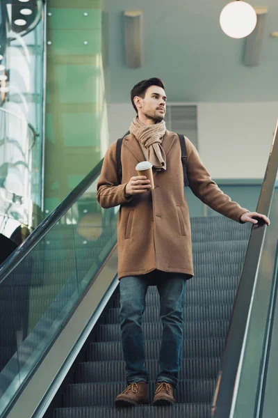 handsome man going down on escalator and holding disposable cup