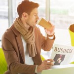 Handsome businessman sitting at table, drinking coffee from paper cup and reading business newspaper in cafe