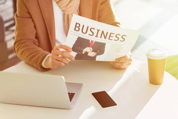 cropped view of man reading business newspaper and sitting at table with laptop, smartphone and disposable cup
