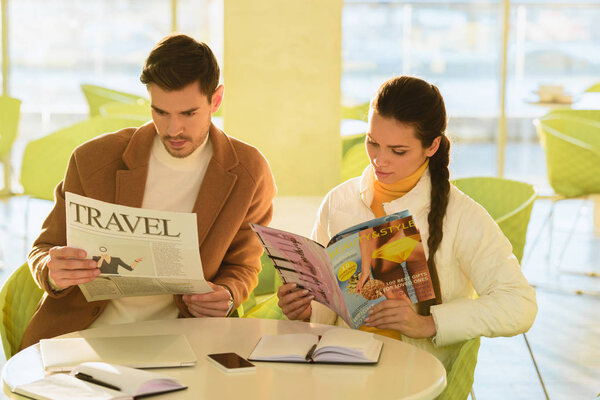 Красивый мужчина читает туристическую газету, а улыбающаяся девушка читает журнал в кафе
