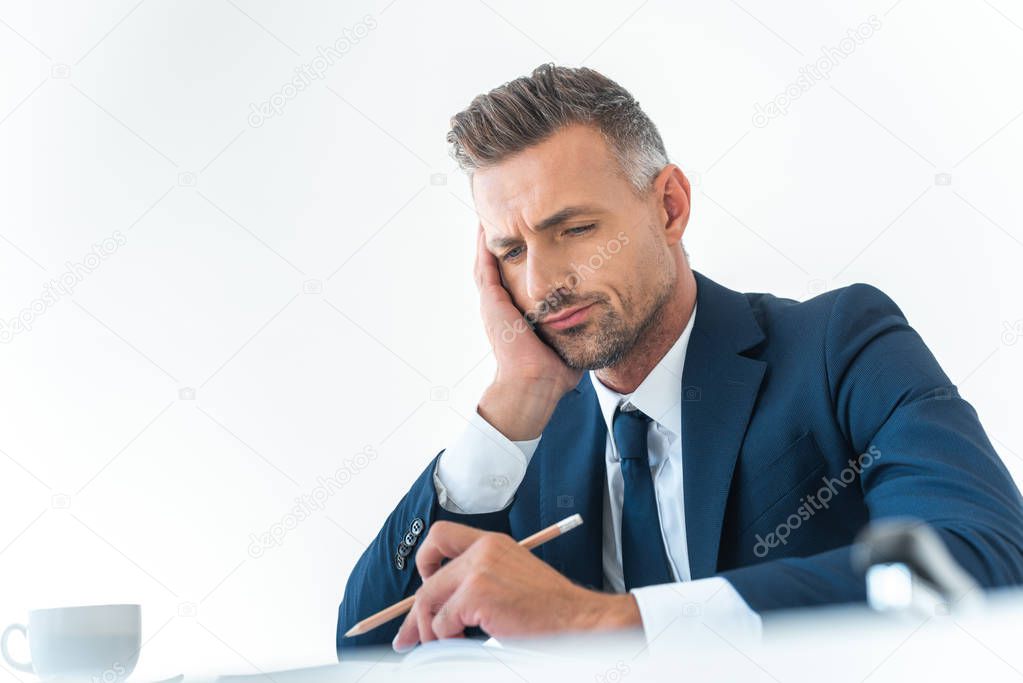 low angle view of tired businessman holding pencil and looking down at table isolated on white