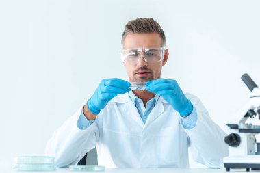 handsome scientist in protective glasses looking at reagent during experiment isolated on white