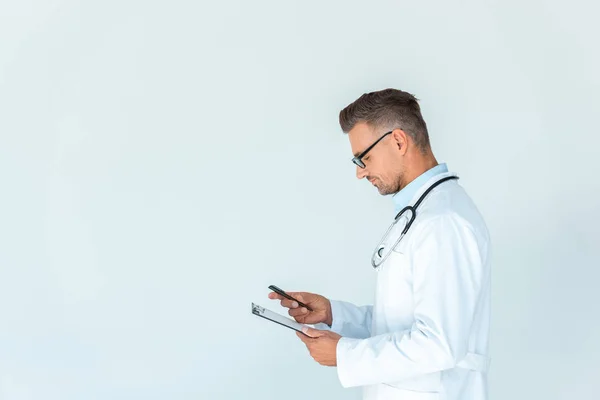 英俊的医生的侧视图在眼镜与听诊器在肩膀举行剪贴板和智能手机查出在白色 — 图库照片