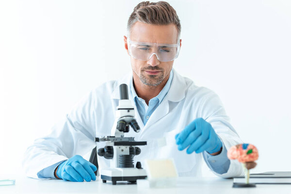 selective focus of handsome scientist in protective glasses making experiment with microscope isolated on white