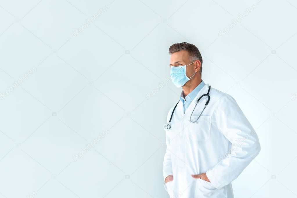 handsome doctor in white coat and medical mask looking away isolated on white