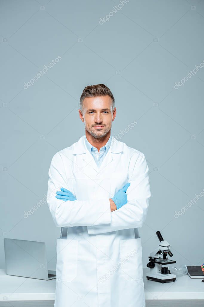 handsome scientist in white coat and latex gloves standing with crossed arms and looking at camera isolated on white