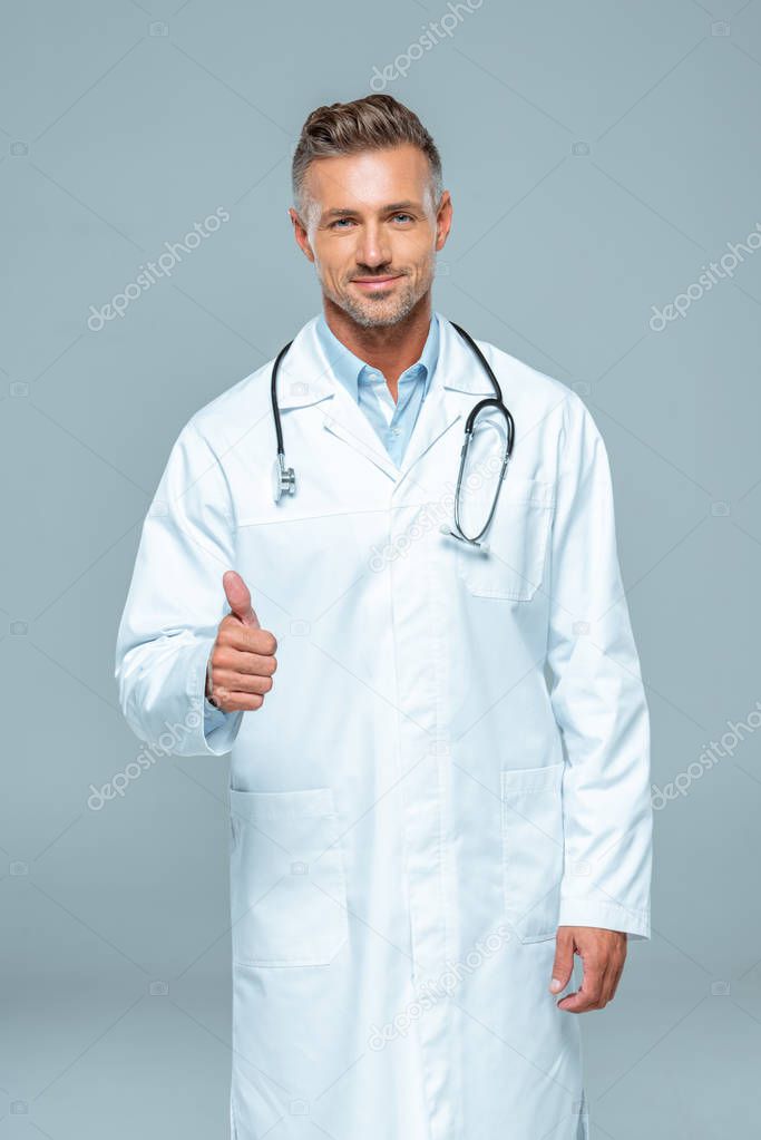 handsome doctor with stethoscope showing thumb up and looking at camera isolated on white