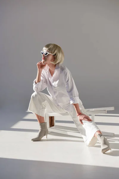 迷人的金发碧眼的女人在太阳镜和时尚的白色装备坐在椅子上 看着白色 — 免费的图库照片