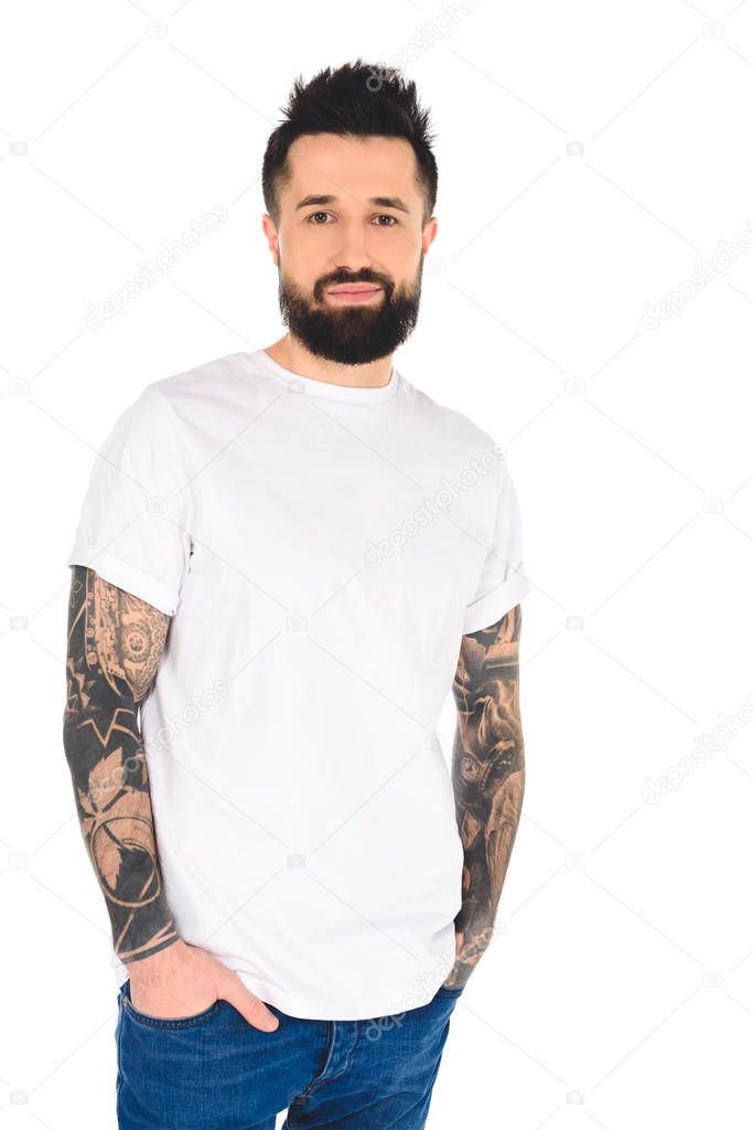 Handsome bearded man with tattoos and hands in pockets looking at camera isolated on white