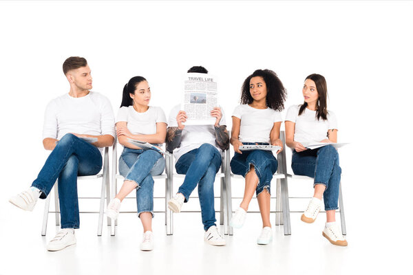 multiethnic group of young people sitting on chairs with crossed legs and looking at man with newspaper and obscure face isolated on white