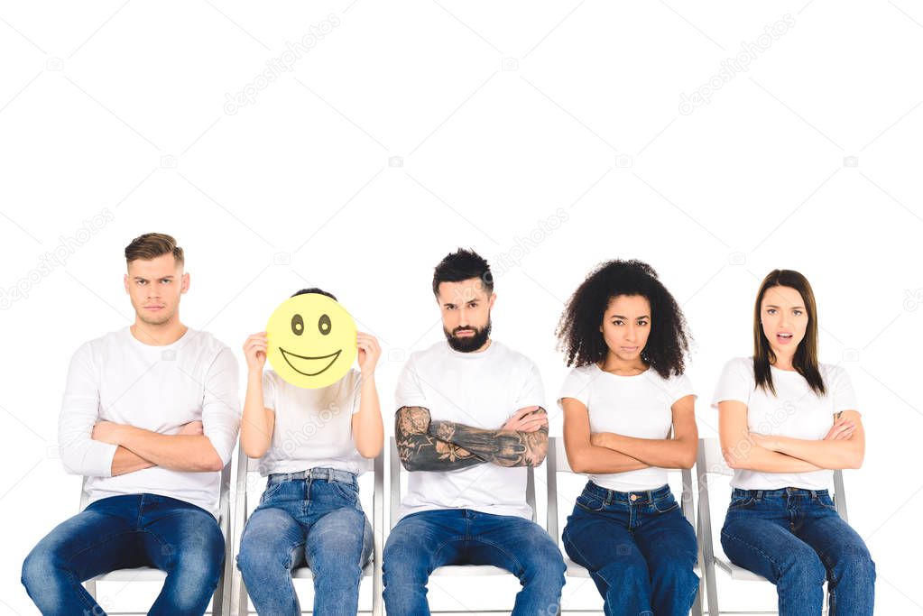 dissatisfied multicultural friends sitting on chairs with crossed arms while girl holding smiling sign isolated on white