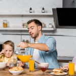 Bonita mujer y lindo chico mirando guapo hombre verter jugo de naranja en vidrio en la cocina